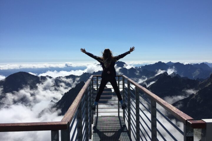 person standing on hand rails with arms wide open facing the mountains and clouds